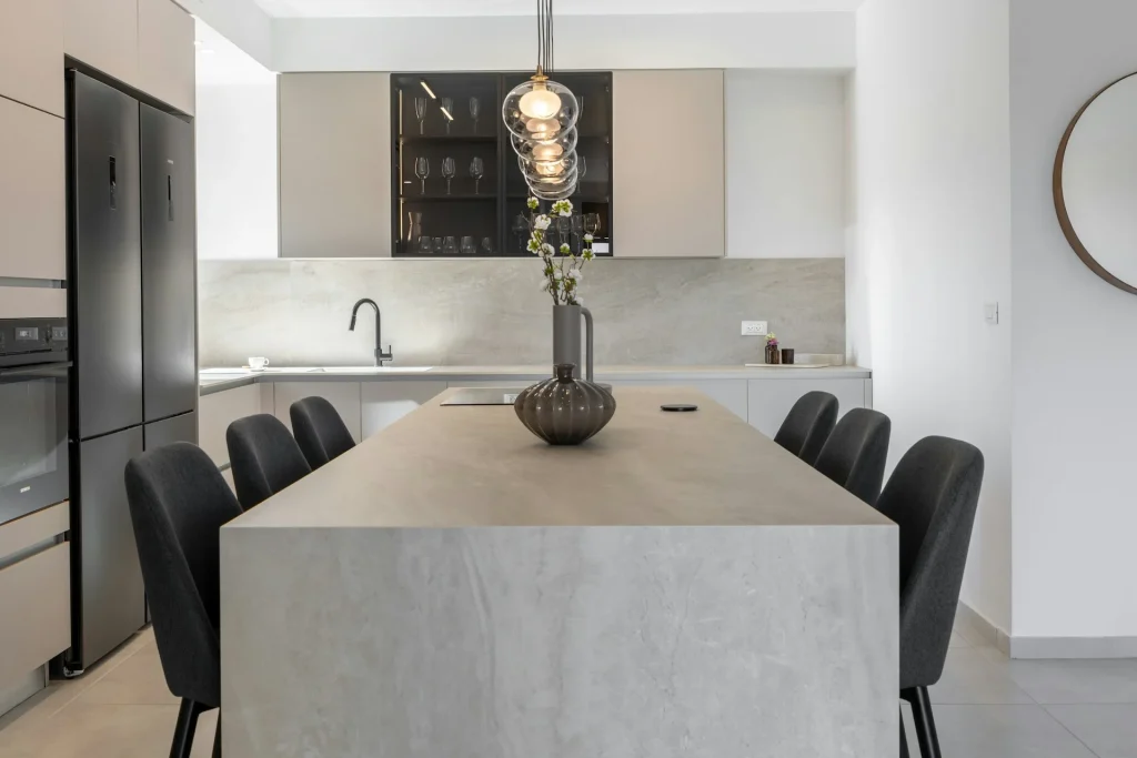 ventajas de las encimeras de microcemento. Gran isla de cocina central con encimera y acabado continuo de microcemento gris claro, rodeada de sillas oscuras y elementos decorativos modernos