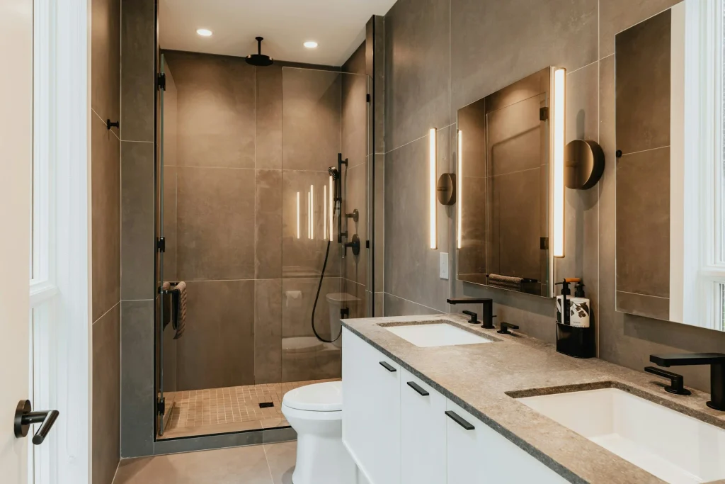 Baño moderno con mampara fija de cristal transparente y perfilería negra, acompañado de un mueble de lavabo doble con encimera de piedra y grifería oscura