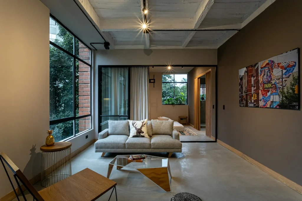Salón de estilo industrial con suelo de microcemento pulido, techo de hormigón visto, pared de ladrillo y amplios ventanales.