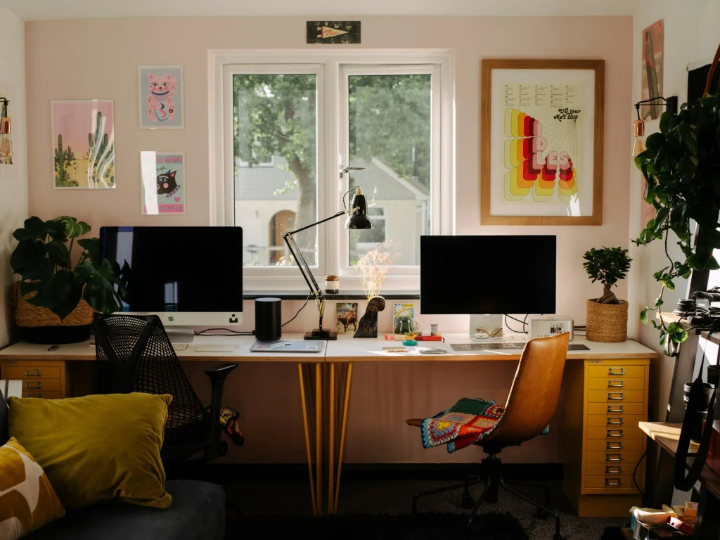 Espacio de trabajo doble en una habitación adaptada como despacho, con un escritorio largo bajo la ventana principal, dos monitores de ordenador, sillas ergonómicas, cajoneras amarillas y cuadros decorativos en paredes de tono rosado tenue.